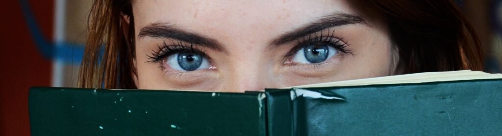 Close-up of a woman with blue eyes peeking over a book, portraying curiosity.